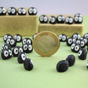 May include: A collection of black and white fuzzy round figurines with large eyes, resembling the soot sprites from the movie 'My Neighbor Totoro'. The figurines are scattered on a green surface, with a Euro coin in the center.