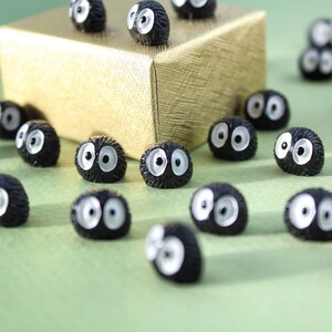May include: A collection of black and white fuzzy round figurines with large eyes. The figurines are scattered on a green surface with a gold gift box in the background.