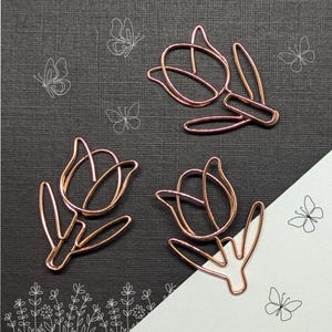 May include: Three rose gold metal paper clips shaped like tulips. The clips are on a black background with white outlines of butterflies.