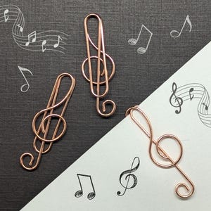 May include: Three rose gold treble clef paper clips on a black and white background. The paper clips are shaped like musical notes.