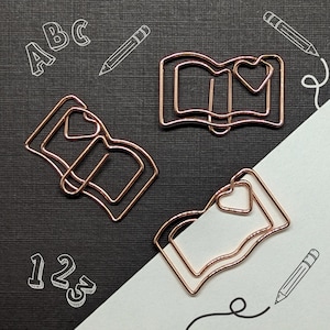 May include: Three gold-toned metal book-shaped paper clips with a heart-shaped design. The paper clips are on a black background with white letters and a pencil drawn on the surface.