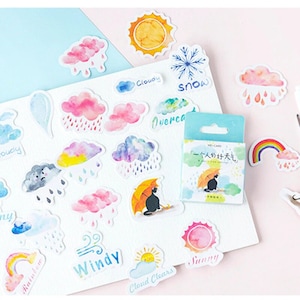 May include: A collection of weather-themed stickers featuring watercolor illustrations of clouds, rain, sun, and rainbows. The stickers include text labels like "Sunny," "Cloudy," and "Windy." A small box of stickers with a cat under an umbrella is also visible.