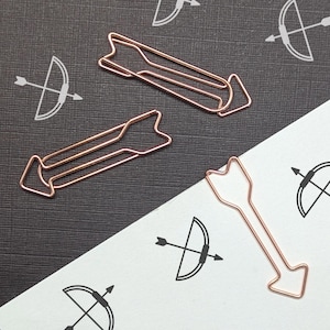 May include: Rose gold arrow-shaped paper clips on a black and white background with bow and arrow illustrations. The paper clips are made of thin wire and have a three-dimensional design. The background is divided diagonally, with a textured black surface on one side and white paper on the other.