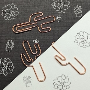 May include: Two rose gold metal cactus-shaped paper clips on a black and white background with a pattern of small cacti and succulents.