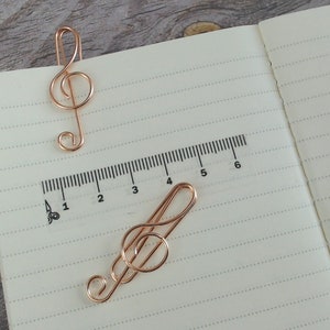 May include: Two gold-coloured metal treble clef bookmarks on a lined notebook page with a ruler printed on the page. The ruler is marked from 1 to 6.