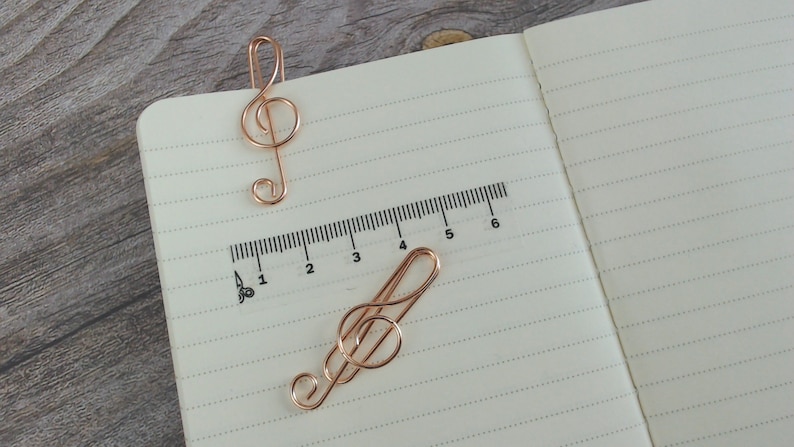 May include: Two gold-coloured metal treble clef bookmarks on a lined notebook page with a ruler printed on the page. The ruler is marked from 1 to 6.