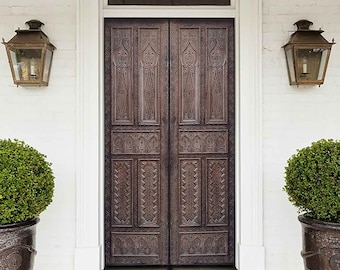 May include: A pair of dark brown wooden double doors with intricate carvings, set in a white brick wall with a white trim. The doors are flanked by two black lanterns with glass panels and a brown woven doormat sits in front of the doors.