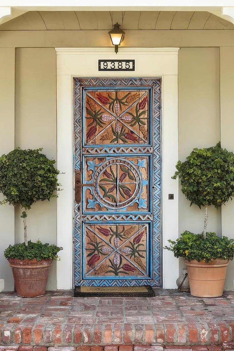 May include: A detailed wooden front door, painted in blue and brown, with carved floral and geometric patterns. The door has the number "9385" above it. Two potted trees sit either side of the door on a brick porch.