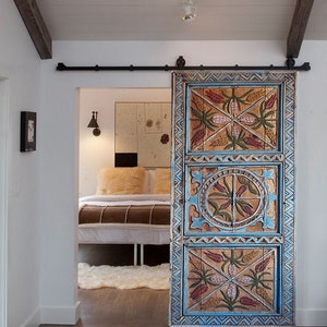 May include: A hand-painted wooden sliding door with a detailed design. The door has a blue border and panels with floral and geometric patterns in red, green, and gold. It is mounted on a black metal track, adding a rustic touch.