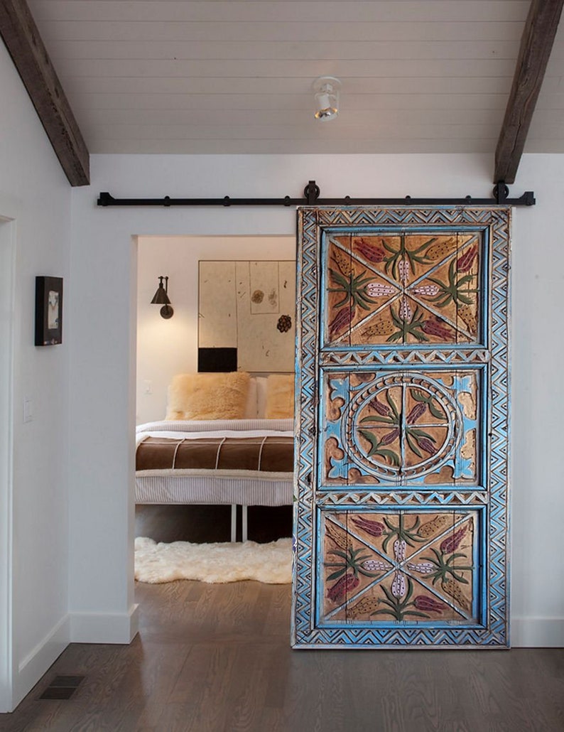 May include: A hand-painted wooden sliding door with a detailed design. The door has a blue border and panels with floral and geometric patterns in red, green, and gold. It is mounted on a black metal track, adding a rustic touch.