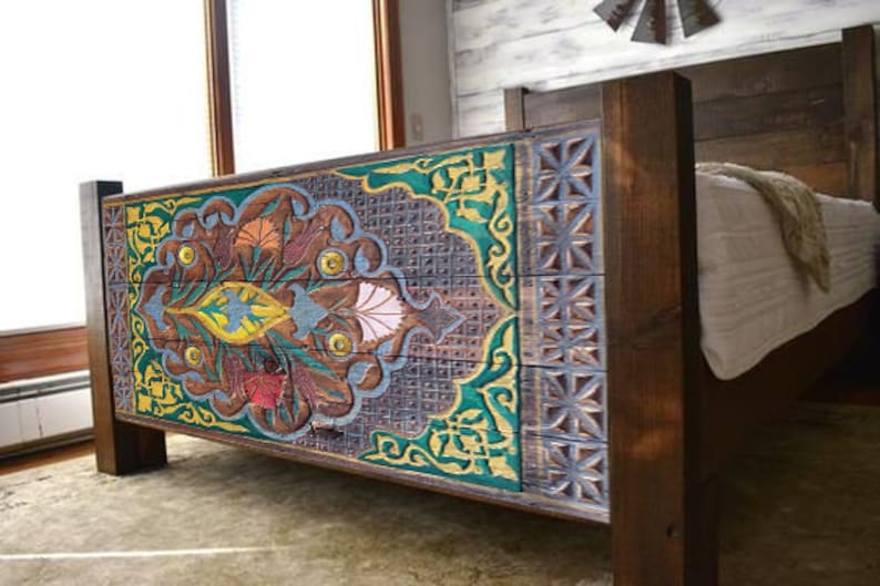 Puede incluir: Una estructura de cama de madera con una cabecera tallada que presenta un dise&ntilde;o floral colorido. La cabecera est&aacute; pintada en tonos de azul, verde, amarillo y rojo. La estructura de la cama est&aacute; hecha de madera oscura y tiene un aspecto r&uacute;stico.