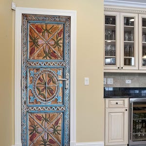 May include: A detailed wooden door with a hand-carved design. The door is painted in shades of blue, orange, and green, with floral and geometric motifs. The door is in a kitchen with cream-colored cabinets and a wine fridge.