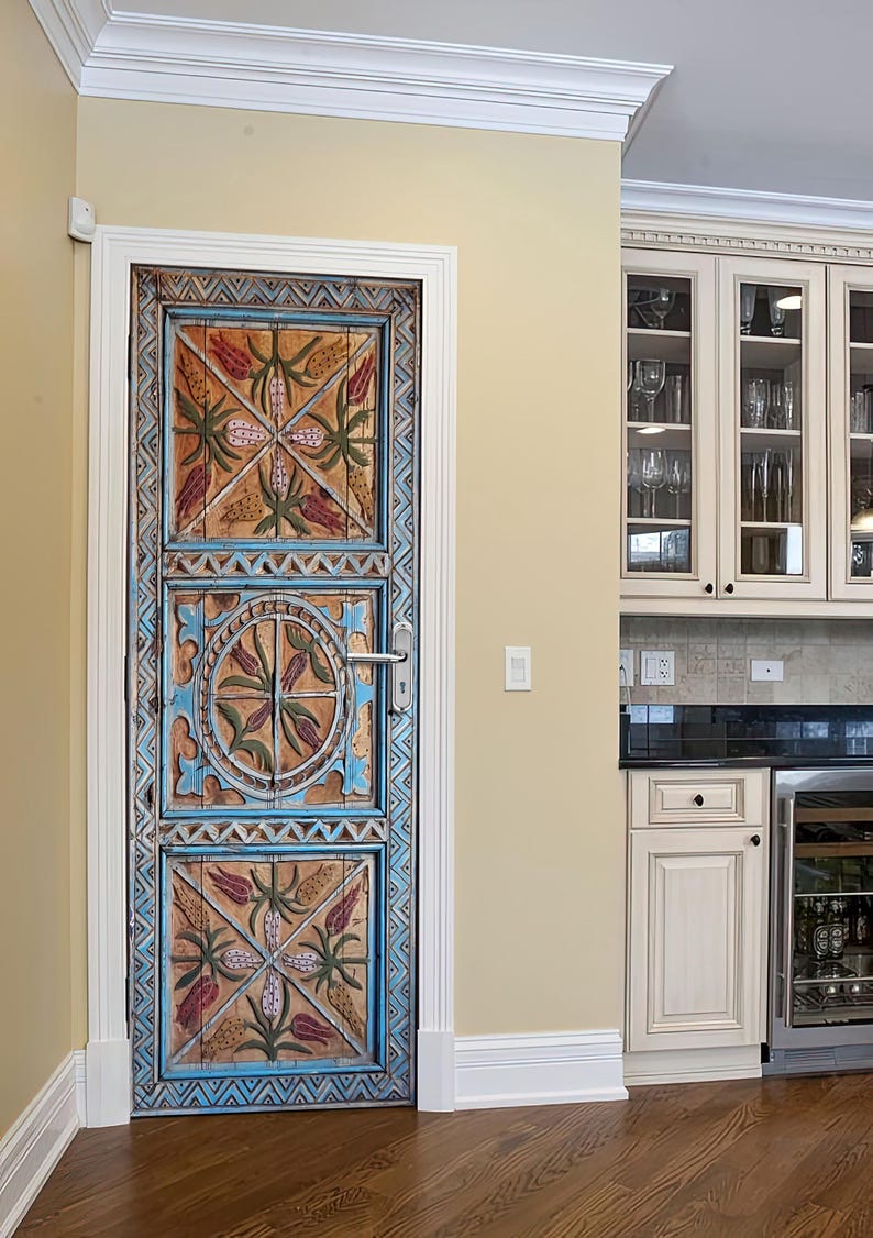 May include: A detailed wooden door with a hand-carved design. The door is painted in shades of blue, orange, and green, with floral and geometric motifs. The door is in a kitchen with cream-colored cabinets and a wine fridge.