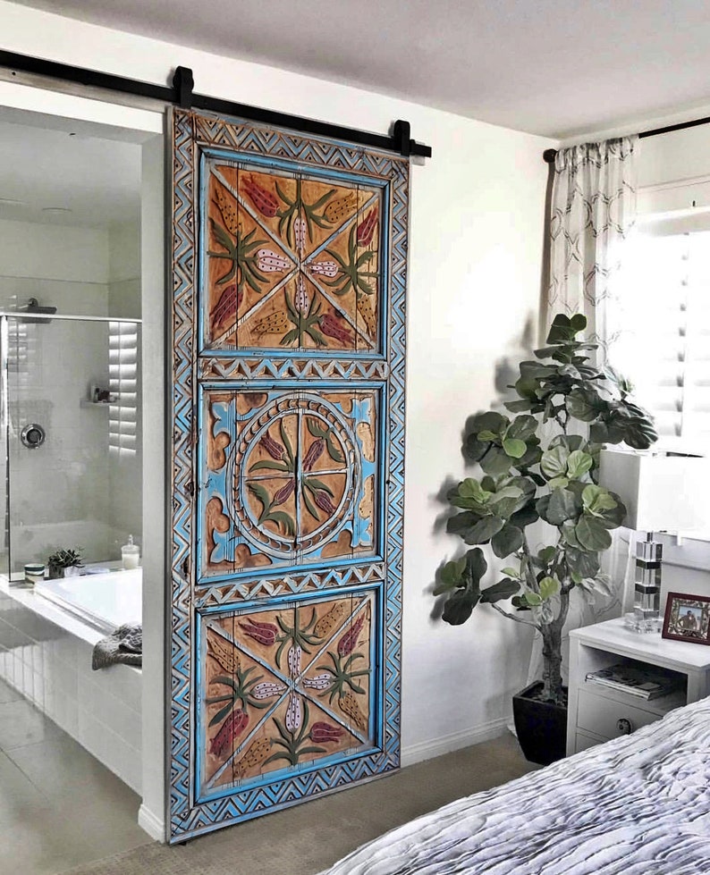 May include: A decorative wooden sliding door with a distressed finish. The door features intricate carvings and is painted in shades of blue, orange, and red. The door is mounted on a black metal track, adding a rustic touch to the room.