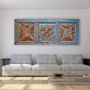 May include: A decorative wooden wall panel with a blue frame and floral patterns in red, brown, and gold. The artwork is displayed above a grey sofa in a modern living room. The room features a large window and wooden flooring.