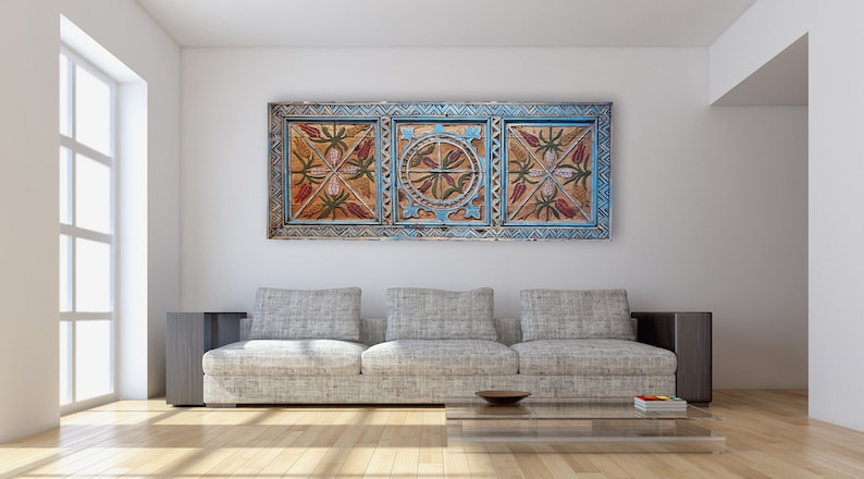 May include: A decorative wooden wall panel with a blue frame and floral patterns in red, brown, and gold. The artwork is displayed above a grey sofa in a modern living room. The room features a large window and wooden flooring.