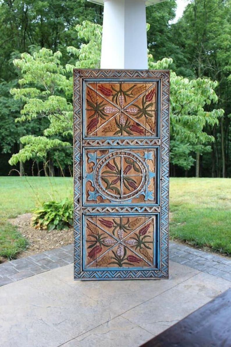 May include: A wooden door with a detailed, hand-painted design. The door features three panels with a repeating floral pattern in red, green, and pink, framed by blue and brown borders. The door is outdoors, set against a backdrop of trees and greenery.