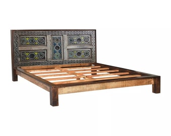 May include: A wooden bed frame with a headboard made from a repurposed door. The door has intricate carvings and is painted in shades of blue, green, and gray. The bed frame is made from dark wood and has a slatted base.