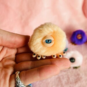 May include: A light orange plush creature with a large blue eye and small black appendages. The creature is held in a hand, with other colourful plush items in the background. The plush has a soft, fuzzy texture.