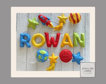 May include: A felt banner with the name "ROWAN" in colorful letters, along with felt stars, planets, and a rocket ship. The banner is strung on a white string and is set against a light wood background. The theme is space and the banner is likely for a child's room.