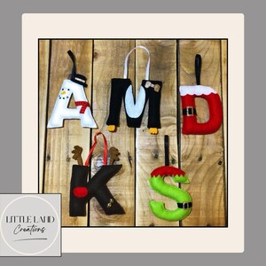 Può includere: Addobbi natalizi in feltro che compongono la parola "AMDKS". Le lettere sono decorate come un pupazzo di neve, un pinguino, Babbo Natale, una renna e un elfo. Gli ornamenti sono bianchi, neri, rossi e verdi, con cordini neri. Sfondo in legno.