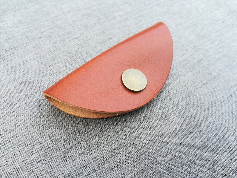 May include: A brown leather key holder with a metal button closure. The holder is shaped like a half-circle.