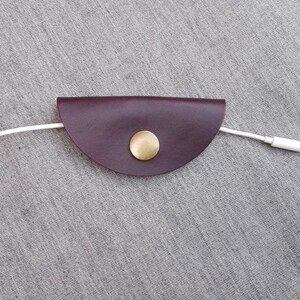 May include: A burgundy leather cord keeper with a gold metal button. A white cord is threaded through the keeper.