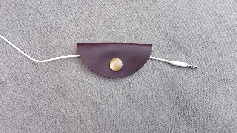 May include: A burgundy leather cord keeper with a gold metal button. A white cord is threaded through the keeper.