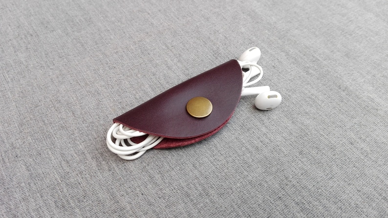 May include: A burgundy leather cord keeper with a gold button, holding white earbuds.