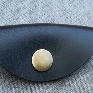 May include: A black leather cord keeper with a brass button. The cord keeper is shaped like a half-circle.