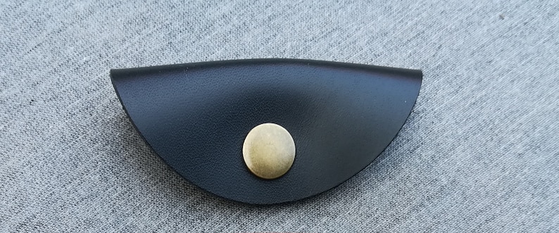 May include: A black leather cord keeper with a brass button. The cord keeper is shaped like a half-circle.