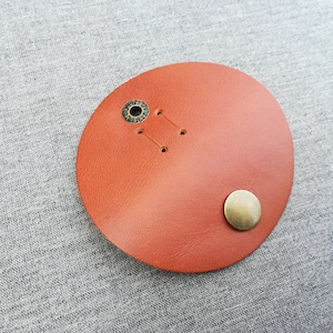 May include: A round, orange leather patch with two metal eyelets and a single metal button.