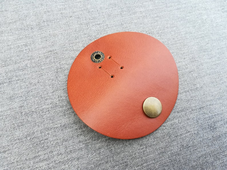 May include: A round, orange leather patch with two metal eyelets and a single metal button.
