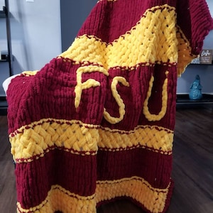May include: A chunky knit blanket in garnet and gold, featuring the letters "FSU" in gold. The blanket has alternating stripes of garnet and gold, with the letters prominently displayed. The blanket is draped over a chair.