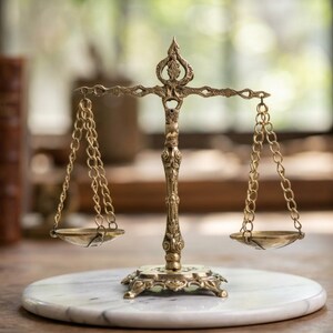 May include: A brass balance scale featuring an ornate central column with intricate scrollwork and a decorative top finial. Two shallow circular pans are suspended by delicate metal chains from a horizontal beam, creating a symmetrical structure.