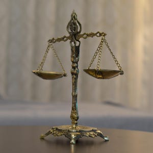 May include: A decorative brass-colored metal scale of justice featuring an ornate central pillar, a detailed base, and two small hanging pans suspended by thin metal chains from a horizontal crossbar with a decorative finial at the very top.