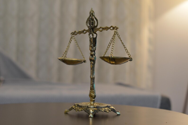 May include: A decorative brass-colored metal scale of justice featuring an ornate central pillar, a detailed base, and two small hanging pans suspended by thin metal chains from a horizontal crossbar with a decorative finial at the very top.