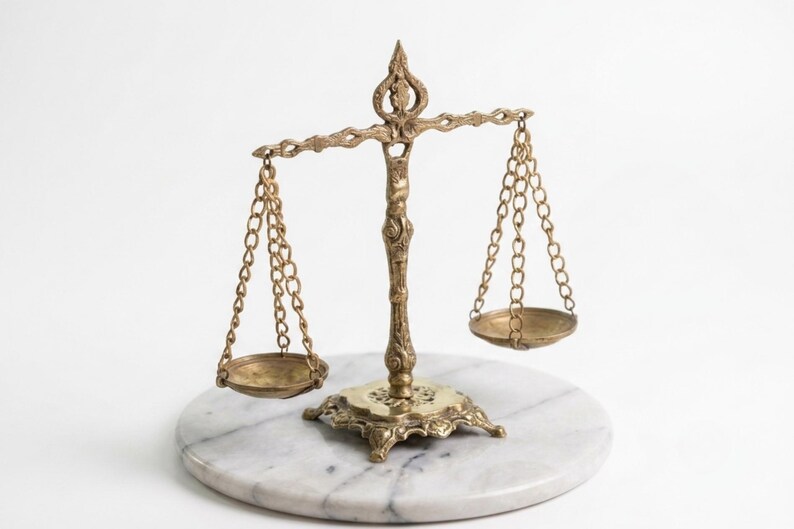 May include: A brass balance scale featuring an ornate central pillar and decorative base. Two circular pans are suspended by thin metal chains from a horizontal beam. The scale is constructed with intricate metalwork details throughout the frame.