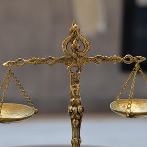 May include: A gold-colored metal balance scale featuring an ornate central pillar with intricate decorative carvings. The scale has two shallow circular pans suspended by thin metal chains from a horizontal beam with detailed filigree patterns.