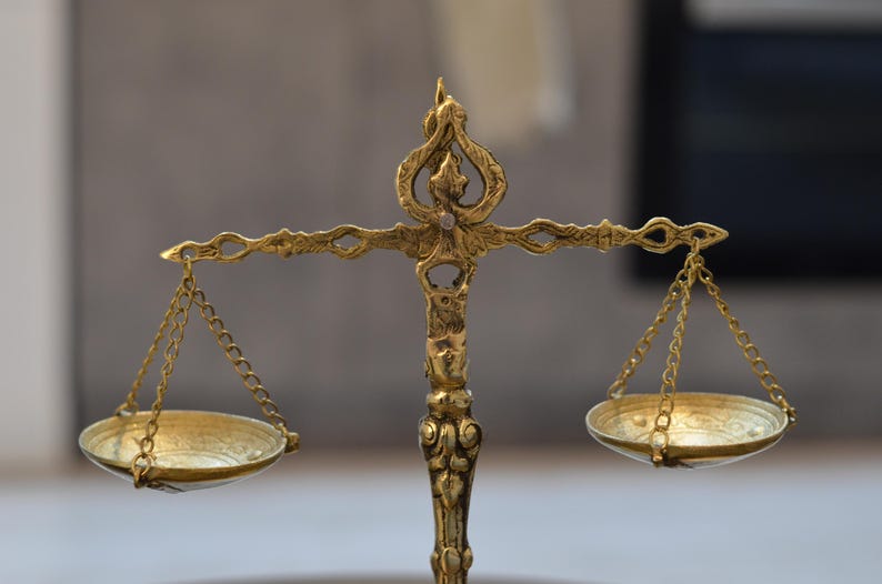 May include: A gold-colored metal balance scale featuring an ornate central pillar with intricate decorative carvings. The scale has two shallow circular pans suspended by thin metal chains from a horizontal beam with detailed filigree patterns.