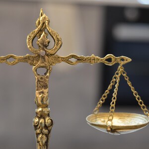 May include: A gold-colored metal balance scale featuring an ornate, decorative central pillar with intricate engravings. The horizontal beam supports two small, shallow circular pans suspended by thin metal chains on each side of the central post.