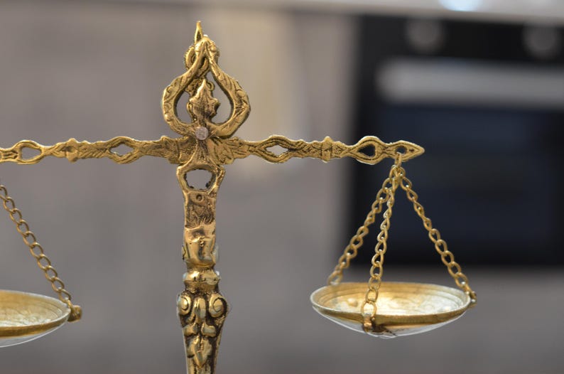 May include: A gold-colored metal balance scale featuring an ornate, decorative central pillar with intricate engravings. The horizontal beam supports two small, shallow circular pans suspended by thin metal chains on each side of the central post.