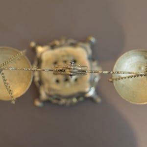 May include: A top-down view of a brass balance scale featuring a central ornate square base, a horizontal beam, and two circular pans suspended by thin metal chains. The metallic structure displays a polished gold-toned finish throughout the object.