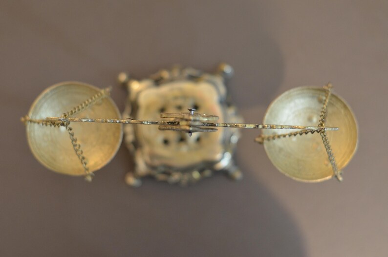 May include: A top-down view of a brass balance scale featuring a central ornate square base, a horizontal beam, and two circular pans suspended by thin metal chains. The metallic structure displays a polished gold-toned finish throughout the object.