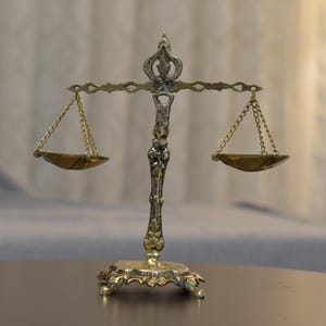 May include: A metallic gold-toned scale of justice featuring an ornate central pillar with decorative scrollwork, a horizontal balance beam, and two small hanging pans suspended by delicate chains on each side, resting on a wide, detailed base.
