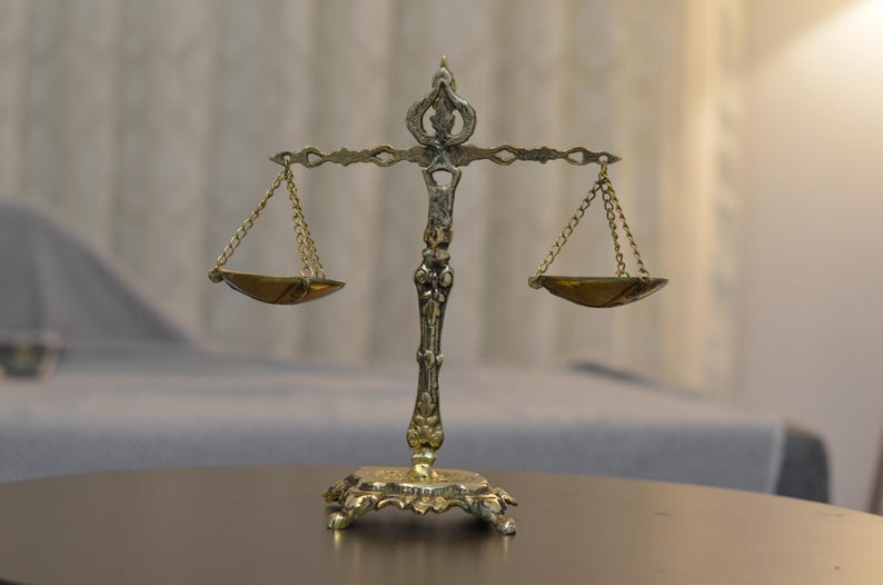 May include: A metallic gold-toned scale of justice featuring an ornate central pillar with decorative scrollwork, a horizontal balance beam, and two small hanging pans suspended by delicate chains on each side, resting on a wide, detailed base.