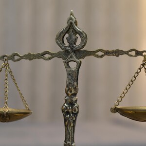 May include: A metallic scale of justice featuring an ornate, textured central pillar with a decorative top finial. A horizontal crossbar supports two small, shallow, circular bowls suspended by thin metal chains on each side of the central column.