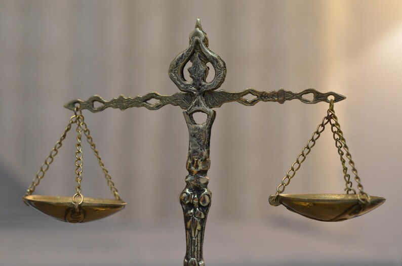 May include: A metallic scale of justice featuring an ornate, textured central pillar with a decorative top finial. A horizontal crossbar supports two small, shallow, circular bowls suspended by thin metal chains on each side of the central column.