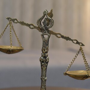 May include: A metallic balance scale featuring an ornate central pillar with decorative carvings. Two shallow, circular pans are suspended from the horizontal beam by thin metal chains on each side, creating a symmetrical weighing instrument.