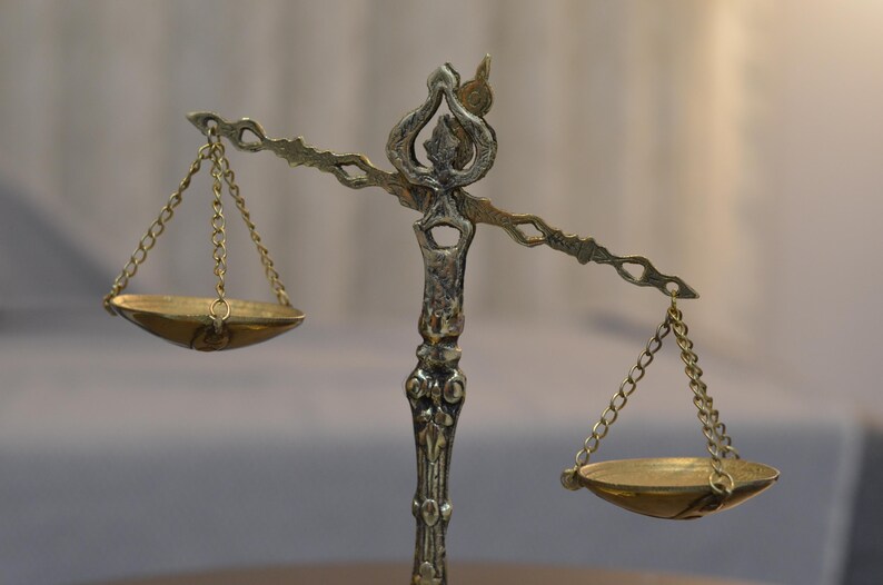 May include: A metallic balance scale featuring an ornate central pillar with decorative carvings. Two shallow, circular pans are suspended from the horizontal beam by thin metal chains on each side, creating a symmetrical weighing instrument.